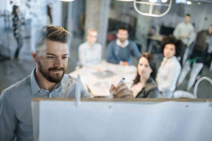 German course in a company. The teacher is writing on a flip-chart, the course participants are sitting around a table.