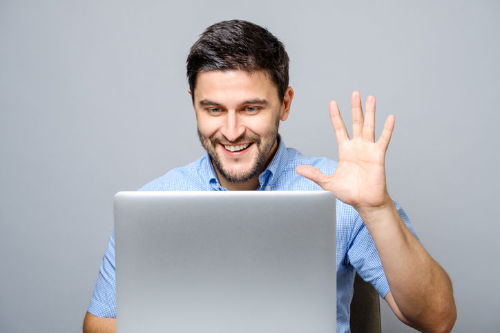 A young man is learning Spanish online via video conference and is waving at his teacher.