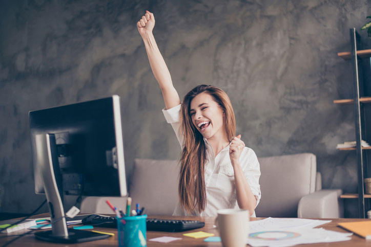 A young woman is sitting in fromt of her computer raising her arms because she has just passed the Spanish exam DELE