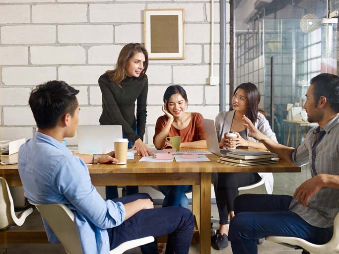 A Swiss-German course in a company. The teacher is standing close to the students, who are sitting around a table.  